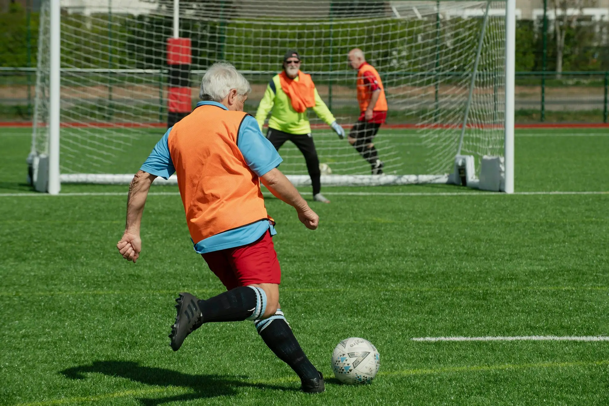 Players in bibs mid-game on a neighbourhood pitch, one striking the ball towards goal