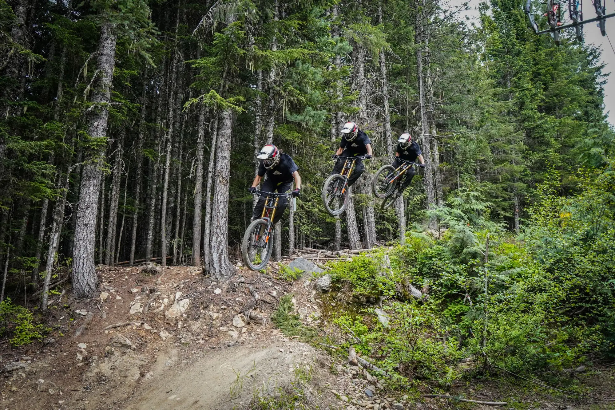 Three mountain bikers airborne over a jump on a forest trail