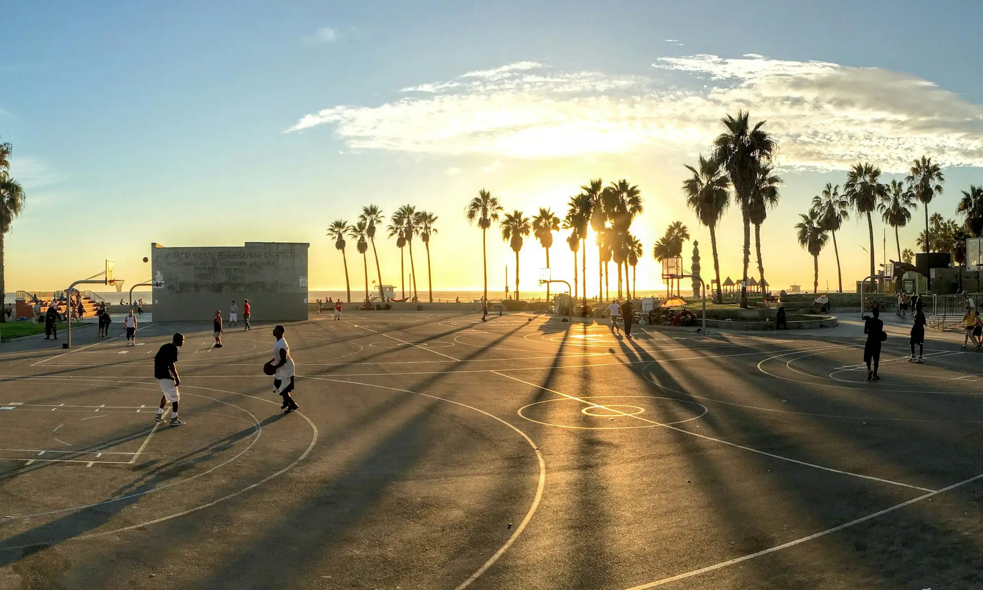 People playing basketball on beachside courts at golden hour — a place a neighbourhood returns to